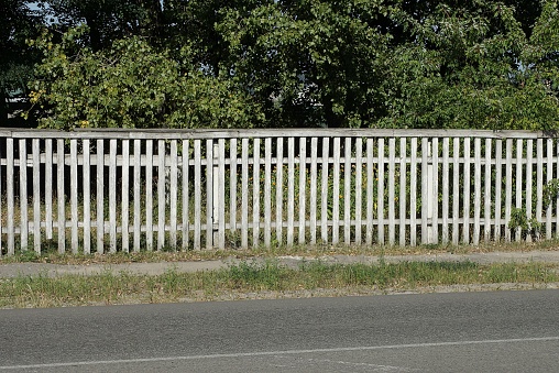 a long white wooden fence in front of green vegetation on a street by a gray asphalt road