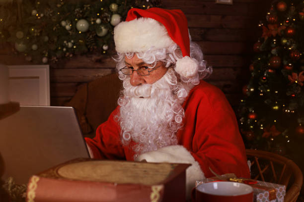 istock Santa Claus using laptop computer sitting in cozy room near fireplace. 1272074184