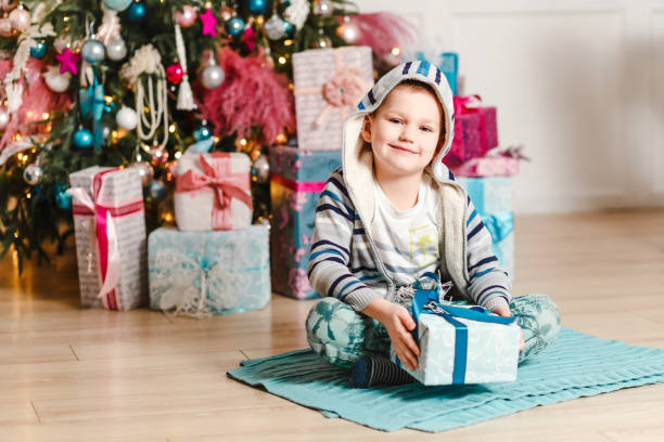 Happy boy sits on plaid under Christmas tree with a gift in his hands