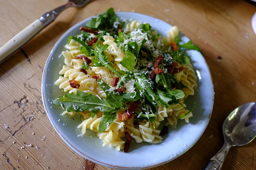 Noodle, arugula, bacon salad topped with parmesan on blue plate.