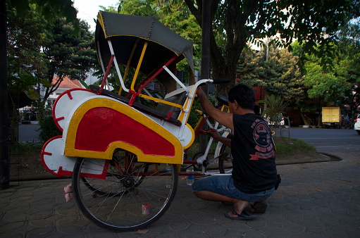 Rickshaw driver in Surakarta, Indonesia, doing some running repairs, on Sept 15 2014. Surakarta, or Solo, is a city in Indonesia which is a large country in Southeast Asia. A rickshaw is a mode of transport requiring a driver. Drivers often repair their own vehicles, which cannot function as modes of transportation unless repaired first when repair is necessary.