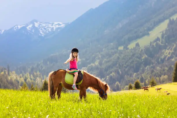 Kids riding pony in the Alps mountains. Family spring vacation on horse ranch in Austria, Tirol. Children ride horses. Kid taking care of animal. Child and pet. Little girl in blooming meadow.