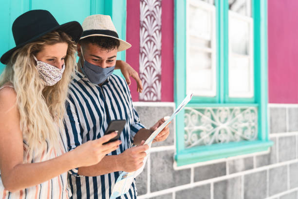 istock Young couple wearing face protective mask while using mobile phone and holding travel map - Travelers people enjoying first trip after corona virus outbreak - Travel health and technology concept 1272119359