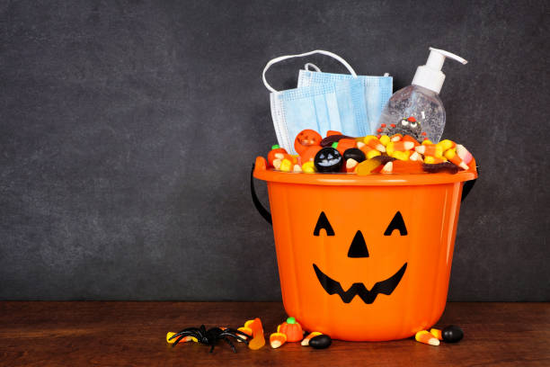 halloween jack o lantern pail with candy and coronavirus prevention supplies against a dark background - halloween color image horizontal side view imagens e fotografias de stock