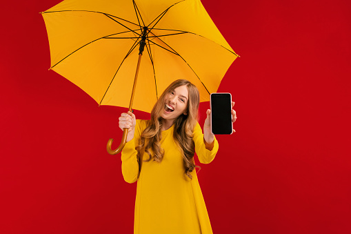 cute young woman in a yellow dress with an umbrella, shows a blank mobile phone screen to copy space on a red background