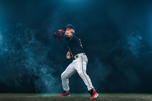 A male baseballs player makes a dramatic play by throwing a ball.  The stadium is blurred behind him. Only the lights of the stadium shine brightly, creating a halo effect around the bulbs.