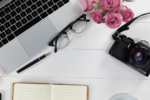 Close up top view of a laptop, a camera, glasses, a notebook and a pen with pink roses arranged on a plain white background