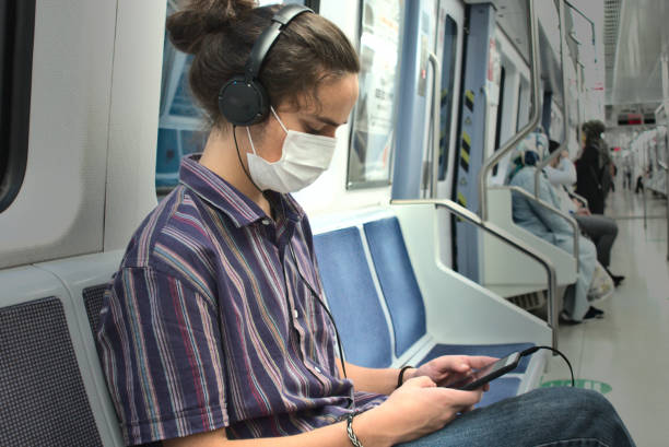 Masked millennial listening to music and playing with his phone inside subway stock photo