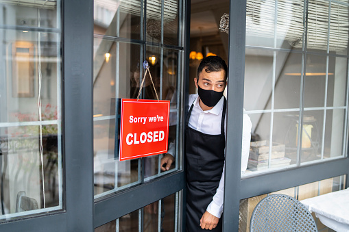 Male restaurant employee wearing protective face mask, watching behind the half-open door where is hanging a signboard 