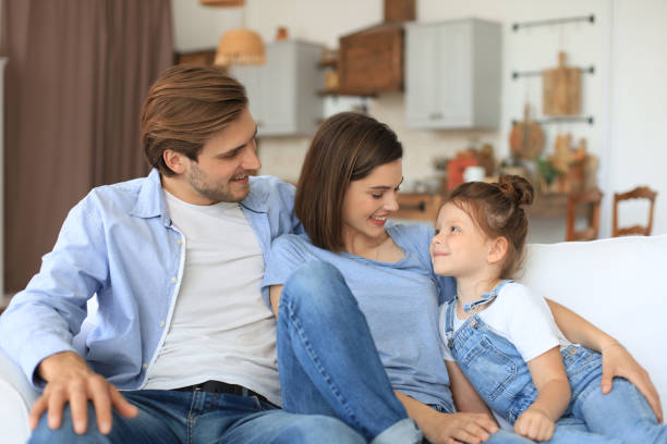 istock Positive friendly young parents with smiling little daughter sitting on sofa together while relaxing at home on weekend. 1272198553