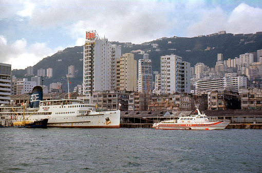 Hong Kong, 1971, The Macau ferry and hydrofoil moored at a wharf, the ferry has a fuel bunker beside it. There are skyscrapers and old low rise buildings side by side, and The Peak is in the background.