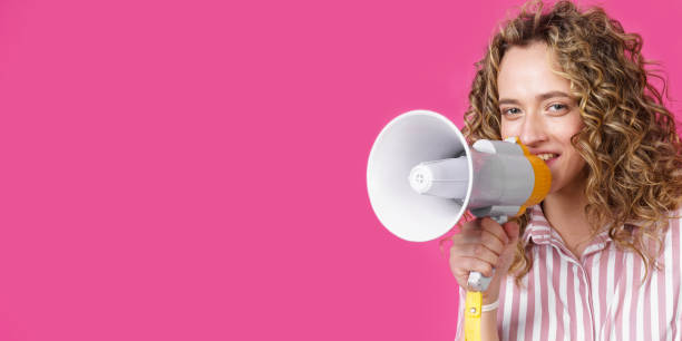 istock Young woman speaks into a megaphone. Isolated pink background. 1272202527