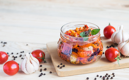 Sun dried tomatoes in glass jar with ripe tomatoes and garlic on cutting board, on wooden background