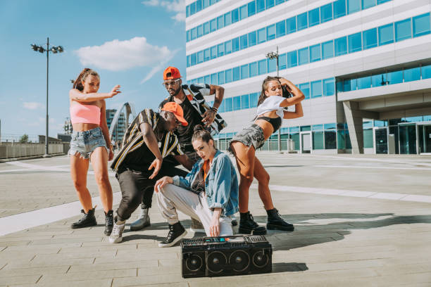 Young man with dreadlocks dances energetically to the rhythm of music in a studio. Expressive and artistic, he moves gracefully with eyes closed, hands expressing the beat. Wearing a smartwatch and wireless earbuds, he listens to his playlist.