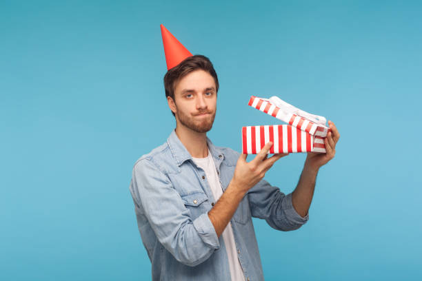 portrait of frustrated man with party cone hat holding opened gift box and looking at camera. studio shot - rudeness ugliness clothing people imagens e fotografias de stock