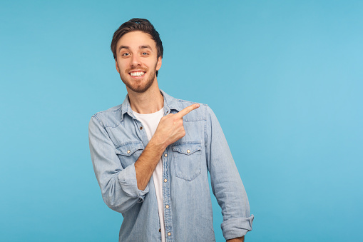 Look at ads here! Portrait of handsome happy man in stylish denim shirt pointing aside, showing blank copy space for idea presentation, commercial text. indoor studio shot isolated on blue background