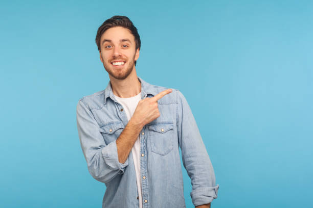 Look at ads here! Portrait of handsome happy man in stylish denim shirt pointing aside, showing copy space Look at ads here! Portrait of handsome happy man in stylish denim shirt pointing aside, showing blank copy space for idea presentation, commercial text. indoor studio shot isolated on blue background pointing stock pictures, royalty-free photos & images