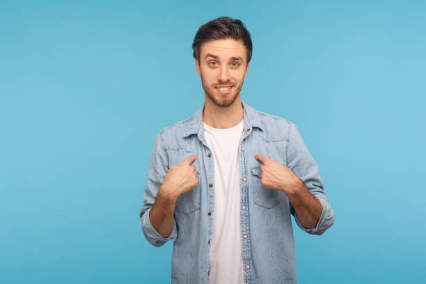 This is me! Portrait of self-confident narcissistic man in worker denim shirt smiling satisfied and pointing himself This is me! Portrait of self-confident narcissistic man in worker denim shirt smiling satisfied and pointing himself, feeling self-important, proud, famous. studio shot isolated on blue background achievement aiming aspirations attitude stock pictures, royalty-free photos & images