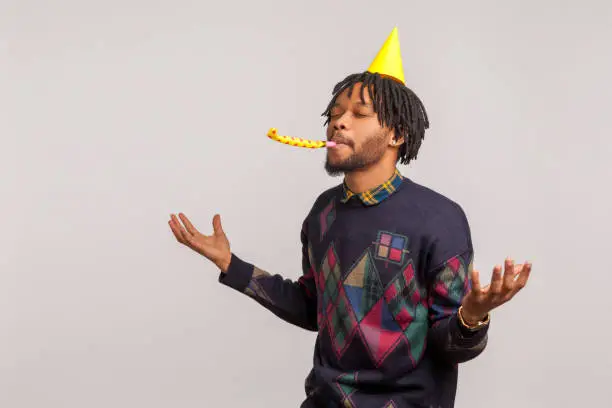 Pleased satisfied african man with dreadlocks in party hat having fun and enjoying blowing party horn with closed eyes, birthday celebration. Indoor studio shot isolated on gray background