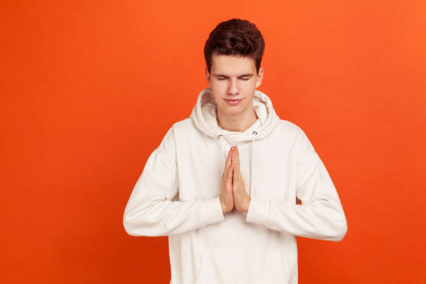 jovem de capuz estilo casual pressionando as mãos juntas mostrando gesto namastê, praticando yoga com rosto pacífico e olhos fechados - peacefull - fotografias e filmes do acervo