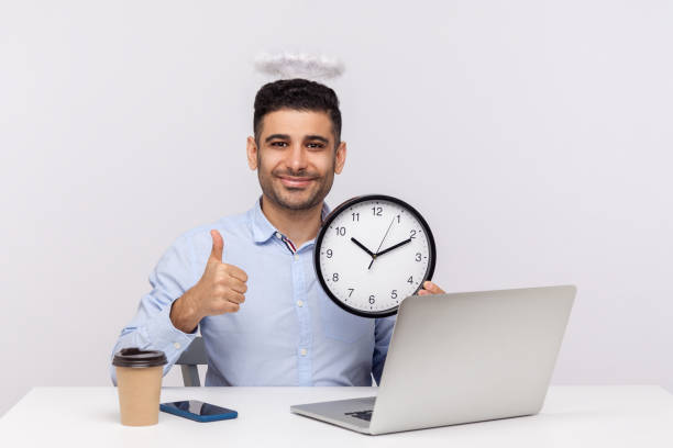 gestión del tiempo. alegre trabajador empleado con nimbus angelical sentado en el lugar de trabajo de la oficina, sosteniendo el reloj - indian culture manager men asian ethnicity fotografías e imágenes de stock