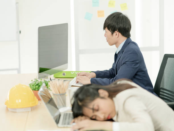 アジア人男性は、スタッフの女性が職場で寝ている間に働いています。過労からストレスを受けたビジネスウーマン - ヌルい職場 ストックフォトと画像