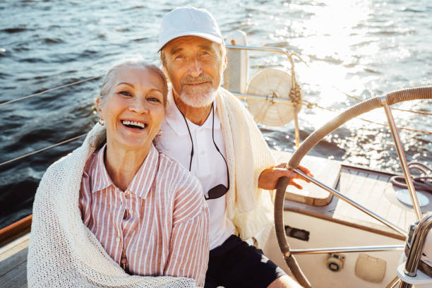 senior couple sitting at the steering wheel on a sailboat and looking at camera - senior adult cheerful adventure discovery imagens e fotografias de stock