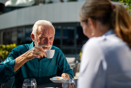 Mature adult businessman drinking coffee in outdoor cafe and looking into pretty young woman