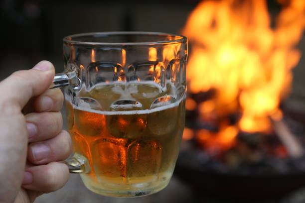 hand holding a lager beer glass with a camp fire in the background. this photo has selective focus. - bonfire beach fire barbecue imagens e fotografias de stock
