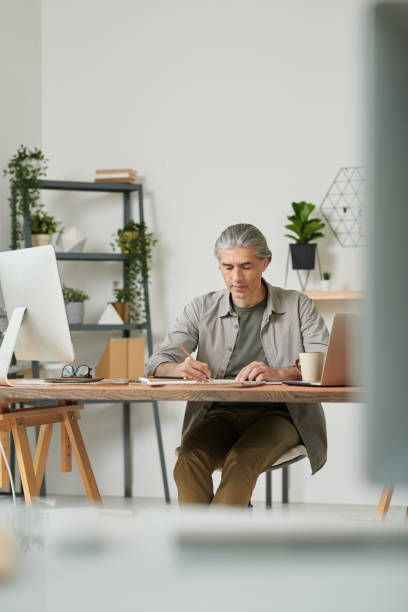 istock Concentrated mature designer sitting at table in modern office and drawing sketch while creating design 1272233058