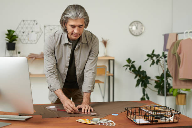 istock Busy mature tailor in casual shirt standing at table and drawing line with ruler while making pattern on brown fabric 1272234215