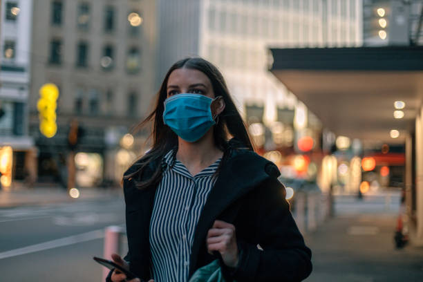 istock Young businesswoman with protective face mask using smartphone while commuting in downtown city street and city buildings with multi-coloured commercial signs at night 1272236420