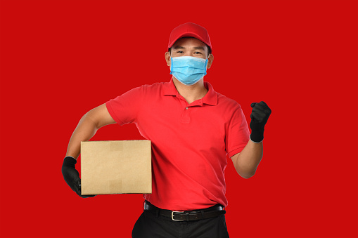 Happy young Asian delivery man in red uniform, medical face mask, protective gloves carry a cardboard box in hands-on red background. The delivery guy give parcel shipment. During COVID-19 outbreak