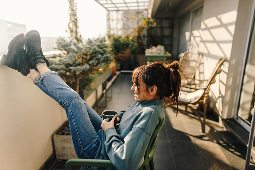 Photo of a woman drinking first-morning coffee on the balcony of her apartment while enjoying early morning sunlight.