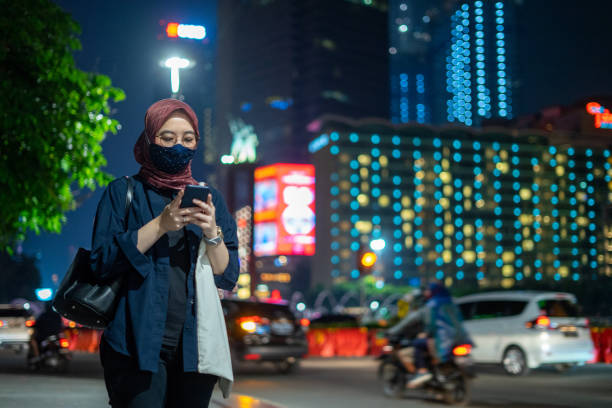 Portrait of Asian Muslim Millennial Business Woman Using her Smartphone in Big Cities at Night After Work Waiting for Public Transportation stock photo
