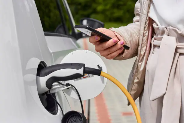 Photo of Young woman charging an electric car at public charging station and pays using a mobile phone. Innovative eco-friendly vehicle.