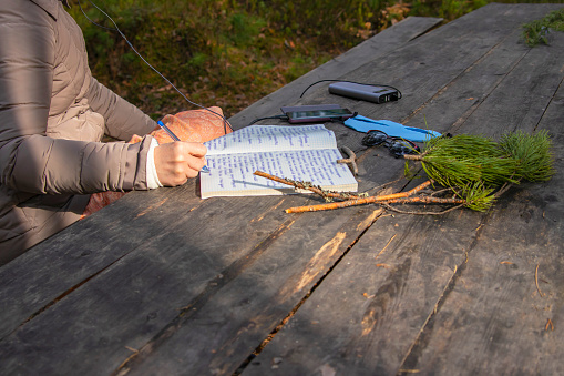 A woman in nature, in a forest, at a wooden table, listens to audio recordings in headphones and writes information in a notebook.
