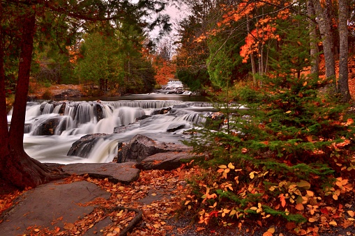 The smaller waterfalls flowing over granite boulders above Bond Falls with the color of autumn. The time exposure evoke feelings of serenity and peace in this autumn forest scenic.  The pathway along the shore is covered with autumn colorful leaves.