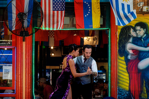Buenos Aires, Argentina - January 24, 2020: Couple dance tango at the entrance of a restaurant in Caminito, in the La Boca neighborhood, with a banner announcing tango lessons