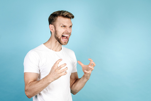 Side view of angry adult man feeling rage, screaming loud, standing isolated on blue copy space background in white t-shirt. Concept of human emotions and facial expressions