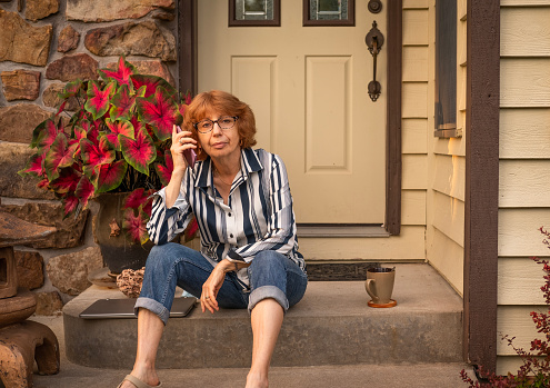 Older woman sitting on front porch of her house talking on the phone in Midwest, USA, in summer;  laptop computer and mug next to her