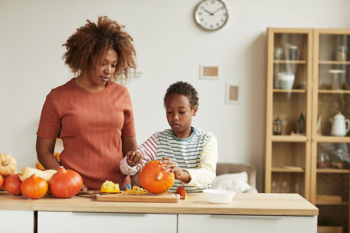 Young adult African American woman wearing casual outfit standing at table watching her child carving pumpkin