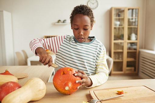 Modern African American boy wearing casual outfit standing at table at home carving face on pumpkin for Halloween party