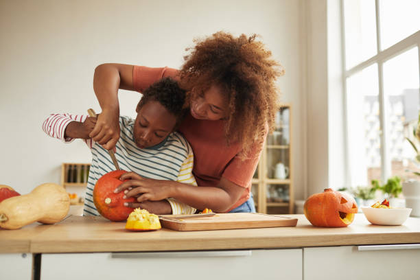 Mom And Son Carving Pumpkin Stylish African American woman spending time with her son standing at table carving pumpkin for Halloween with kitchen knife together pumpkin decorating stock pictures, royalty-free photos & images