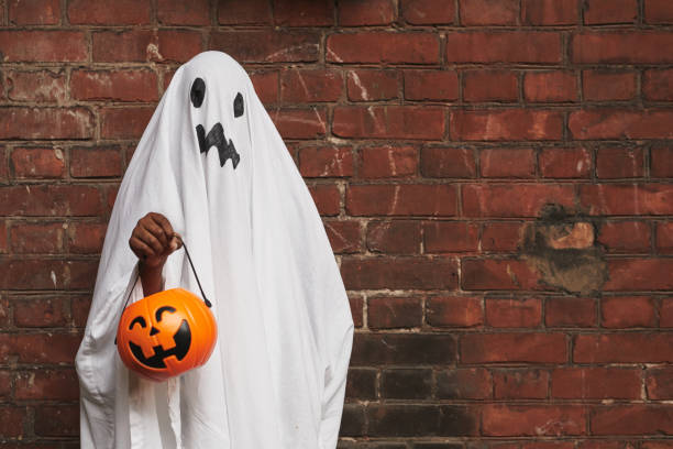 Kid Dressed Up As Ghost Unrecognizable kid dressed up as ghost wearing white fabric holding jack o' lantern basket for sweets standing against brick wall costume stock pictures, royalty-free photos & images