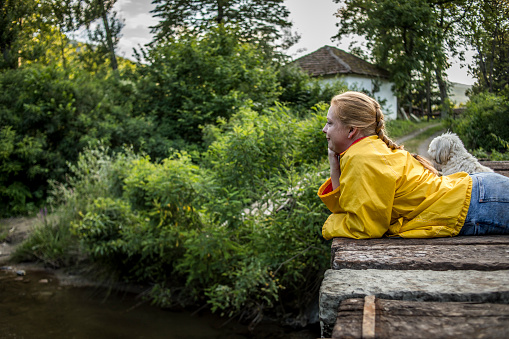 A young woman in a yellow jacket is sitting on the bridge over the river and watching nature - The nature lover enjoys the soothing scenes of nature