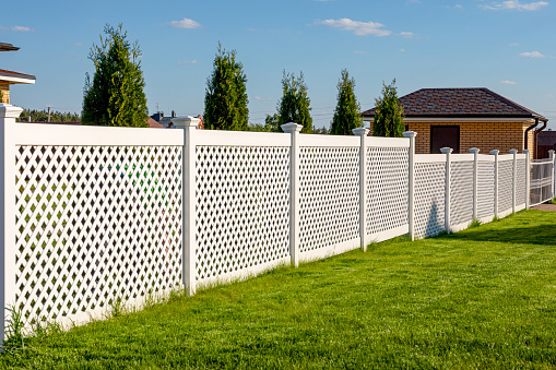 White vinyl fence in a cottage village. Several panels are connected by columns. Tall Thuja bushes behind the fence. Fencing of private property.