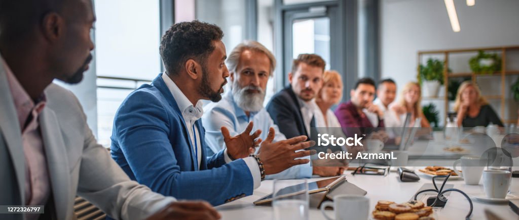 Executive Team Listening to Contrary Views from Colleague Determined African businessman expressing opinions to junior and senior colleagues on management team in conference room. Organized Group Stock Photo