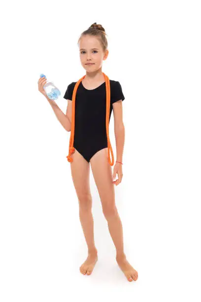 Picture of girl gymnast in black trico in full height with a jumping-rope around her neck and a bottle of water in hand.