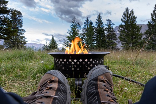 A man is relaxing by a campfire in summer in Banff National Park. He is contemplating the sunset and beautiful mountain range. He is relaxing it is a tranquil scene. It is a cold evening and the fireplace is keeping him warm.